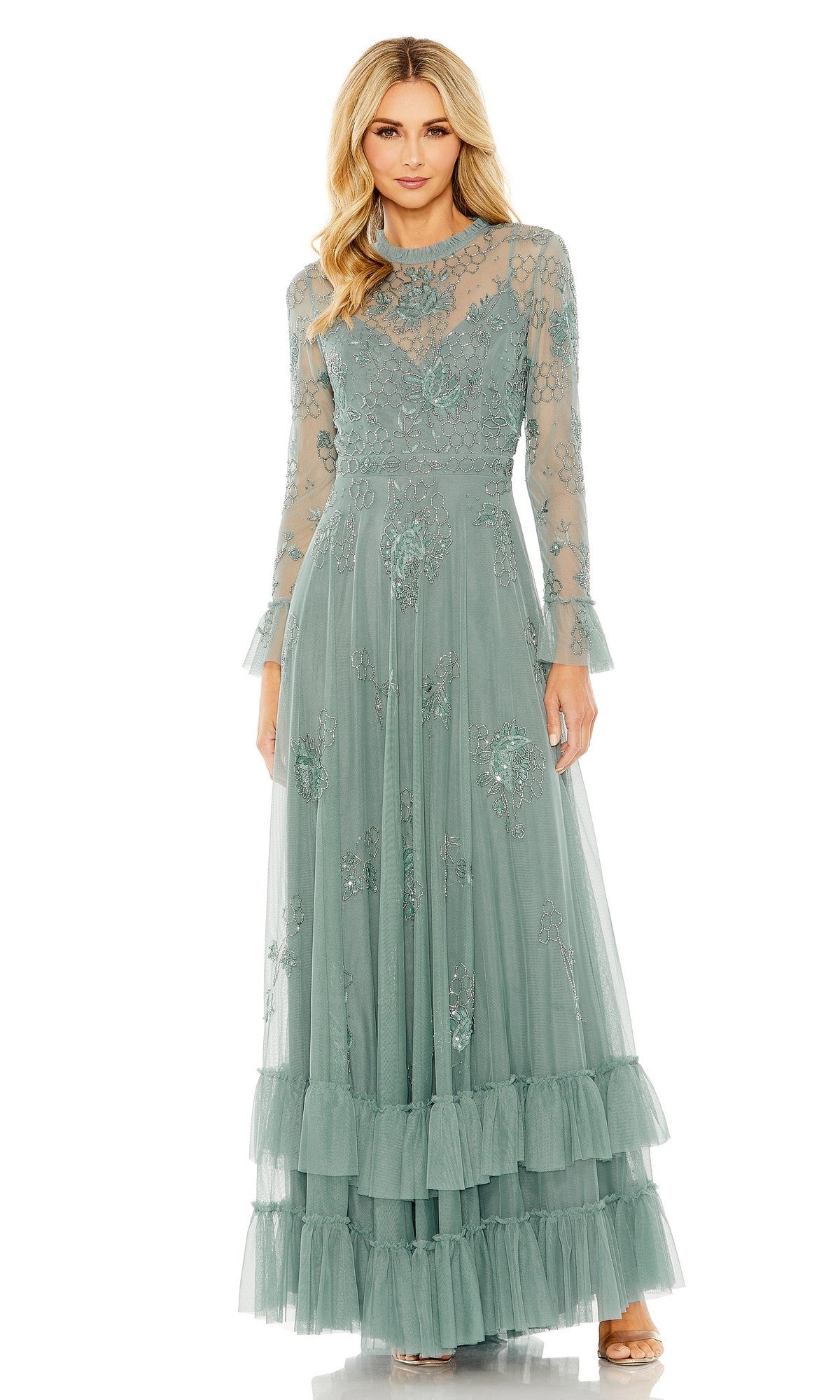 A woman wearing the Mac Duggal 9249 long formal dress in Spruce, featuring sheer long sleeves, beaded floral embellishments, lace detailing, and a ruffled hem, stands against a white background.